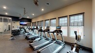 This fitness center offers a comprehensive workout environment across a single floor, designed to accommodate a wide range of exercise routines. The main cardio area is lined with a row of True-brand treadmills and ellipticals positioned along a wall of large windows with roller shades, allowing natural light to fill the space while offering exterior views. Industrial-style ceiling fans and recessed lighting provide consistent climate control and illumination throughout. A wall-mounted flat-screen TV is positioned for viewing during workouts. The free weight section features a full rack of dumbbells alongside a weight bench, supported by mirrored walls and cable machine equipment. A dedicated studio room houses True cycling bikes, colorful kettlebells, medicine balls, and stability balls, offering a versatile space for group or individual training. A separate smaller room provides additional equipment including a treadmill and rowing machine, decorated with a motivational fitness-themed wall graphic. The entry area includes a water station, restrooms, and posted facility rules, establishing a functional and organized welcome zone. Durable carpet flooring throughout and neutral-toned walls with dark accent finishes give the space a clean, modern aesthetic suited to focused, high-quality workouts.