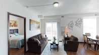 This single-floor space centers around a well-appointed living room that balances comfort and functionality. The room features dark hardwood-style flooring that contrasts cleanly with the white walls, creating a crisp, modern aesthetic. A large sliding glass door dominates one wall, flooding the space with natural light and providing direct access to a private balcony with outdoor views of the surrounding area. The living area is generously proportioned, comfortably accommodating a full-sized sofa and an armchair arranged around a dark wood coffee table atop a plush area rug. Decorative wall art, including a vibrant cityscape canvas, adds a touch of character to the neutral palette. A ceiling-mounted light fixture provides ambient illumination, while a side table lamp offers additional warmth. Vertical air vents integrated into the ceiling suggest a central HVAC system. A doorway visible to the left hints at an adjacent bedroom, indicating a thoughtful, connected layout. Overall, the space presents a clean, contemporary feel with an emphasis on natural light and practical living.