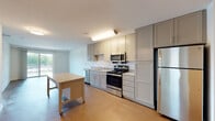 This single-floor unit offers a clean, contemporary design with a neutral color palette throughout. The kitchen serves as a central feature, outfitted with gray shaker-style cabinetry extending to the ceiling for ample storage. Stainless steel appliances include a range, over-the-range microwave, dishwasher, and refrigerator. Granite-style countertops complement the cabinetry, while a mobile island provides additional prep and dining space. The kitchen flows into an open living area finished with wood-look flooring, creating a bright and cohesive common space.

The bedroom areas feature plush wall-to-wall carpeting and large windows that bring in generous natural light and outdoor views of the surrounding greenery. The clean white walls contribute to an airy, open feel.

The bathroom is finished with classic white subway tile surrounding a full soaking tub and shower combination, accented by a decorative tile border. Wood-look tile flooring adds warmth, while a gray vanity with a white countertop and a mirror complete the space. Overall, the unit presents a functional, well-designed layout with consistent modern finishes throughout.