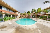 This multi-unit residential complex is arranged around a spacious central courtyard that serves as the heart of the community. The focal point of the outdoor area is a generously sized, kidney-shaped swimming pool with clear turquoise water, surrounded by a wide concrete sun deck equipped with lounge chairs for relaxation. A gated spa area is also present adjacent to the pool, offering an additional amenity for residents.

The buildings frame the courtyard on multiple sides, presenting a two-story layout with exterior walkways and balconies that overlook the pool area. The architecture reflects a Spanish-influenced style, distinguished by classic red-tile roofing along one wing, stucco exterior walls in warm tan tones, and arched covered walkways at ground level. Bold purple accent doors and trim add a distinctive visual character throughout the complex.

Tropical and subtropical landscaping enhances the courtyard setting, with mature palm trees, banana plants, and lush shrubbery providing shade and a resort-like ambiance. Outdoor seating and dining furniture are positioned along the periphery of the deck, offering communal gathering spots. The complex also features secure fencing around the pool area and clearly posted safety signage. Stairways connect the ground level to the upper-floor walkways, and bicycle storage is visible near the building entrance, reflecting a community-oriented residential environment.