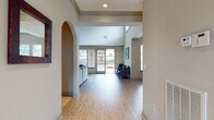 This single-floor clubhouse offers a thoughtfully designed community gathering space that blends open-concept living with functional amenities. The interior greets visitors with a wide, light-filled corridor featuring elegant arched architectural details, light hardwood-style flooring, and soaring ceilings that lend an airy, spacious feel throughout. Crown molding and recessed lighting add refined finishing touches to the neutral-toned walls.

The main lounge area is generously proportioned, anchored by a plush sectional sofa and accent seating, with a decorative mirror and artwork enhancing the welcoming ambiance. Abundant natural light pours in through large windows and glass-paned French doors that open directly to the outdoor pool area, creating a seamless indoor-outdoor connection.

A dedicated dining and kitchenette area provides practical convenience, featuring dark cabinetry, a counter with small appliances, a dining table with seating, and bright windows that keep the space feeling open and comfortable.

Outside, a covered patio with ceiling fans and high-top seating overlooks a sparkling community pool surrounded by mature palm trees and lush landscaping. Brick columns frame the covered outdoor space, adding architectural character. The overall environment is clean, modern, and inviting — ideal for social gatherings and everyday community use.