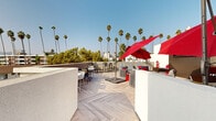 Located at 1558 Gordon St in the heart of Hollywood, Los Angeles, this rooftop terrace offers an inviting open-air retreat with a distinctly modern aesthetic. The deck is finished in a light-toned herringbone-patterned wood-look flooring that lends a clean, sophisticated feel underfoot throughout the entire space.

The terrace is thoughtfully organized into several distinct zones. A sunbathing area features striped black-and-white chaise lounges accented with red throw pillows, shaded by bold red cantilever umbrellas that serve as striking visual anchors across the deck. A built-in bench seat with black-and-white striped cushions and vibrant red accent lanterns provides a cozy nook ideal for relaxing conversation.

Multiple outdoor dining and seating arrangements are positioned across the terrace, including a teak-style dining table with slatted chairs and a casual lounge grouping with wicker and upholstered seating. An outdoor flat-screen television is mounted on one of the white stucco architectural walls, adding an entertainment dimension to the space.

Surrounded by low white parapet walls, the terrace opens to panoramic views of towering Los Angeles palm trees and the surrounding Hollywood skyline under wide-open blue skies, capturing the quintessential Southern California outdoor living experience.