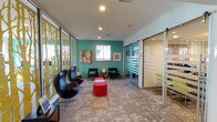 This contemporary single-floor space is thoughtfully designed to create a welcoming and visually dynamic environment. The entry area greets visitors with a striking hexagonal tile floor that transitions into plush, patterned carpet throughout the interior. Just inside the entrance, a self-serve refreshment station is neatly integrated into the design, featuring a coffee machine, mini refrigerator, and bold geometric accent wallpaper framed by warm wood paneling and a large display screen.

The main lounge area is defined by bold color choices, including a standout teal accent wall adorned with curated artwork and framed prints. Iconic egg-style black chairs, classic armchairs, and vibrant red ottoman stools furnish the space, creating an inviting and modern atmosphere. Decorative gold tree-motif panels serve as a striking visual divider, adding an artistic, nature-inspired element to the layout.

Adjacent to the lounge, glass-walled rooms enclosed by sliding frosted-glass panel doors provide private meeting or office spaces, offering transparency while maintaining functionality. A workstation area with computer terminals and colorful accent furnishings rounds out the space, supporting both collaborative and individual tasks. Natural light filters in through multiple windows, enhancing the overall bright and energetic ambiance of this well-appointed space.