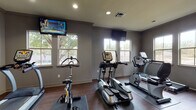 This single-floor fitness center is thoughtfully designed to support a full range of workout routines. The facility is divided into several distinct zones, each tailored to a specific type of training. The cardio area is generously sized and outfitted with treadmills, elliptical machines, a stationary bike, and a recumbent bike, all arranged to take advantage of natural light streaming in through multiple windows. Wall-mounted televisions keep users entertained during their workouts, while ceiling fans help maintain airflow and comfort throughout the space. The strength training zone features a comprehensive set of dumbbells neatly organized on tiered racks, accompanied by benches for a variety of exercises. Full-length mirrors line the walls, enhancing the sense of space and allowing users to monitor their form. A dedicated functional training area offers a carpeted floor, stability balls, medicine balls, and a multi-purpose rack, providing an ideal environment for stretching and dynamic workouts. A Hoist multi-function cable machine adds further versatility to the strength section. A built-in refreshment station with dark cabinetry, granite-style countertops, and a water cooler offers a convenient amenity. Rich hardwood-style flooring and neutral gray walls give the entire facility a modern, clean aesthetic throughout.