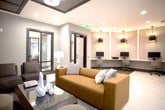 Located at 4558 Roswell Rd NE in Sandy Springs, GA, this single-floor amenity space presents a thoughtfully designed environment with a cohesive contemporary aesthetic throughout. The layout encompasses several distinct functional areas unified by a consistent palette of gray tones, dark tile flooring runners, and light-colored accent surfaces.

The business center features a long built-in desk accommodating multiple workstations, complemented by dramatic pendant lighting and striking blue LED accent columns that lend a modern, energetic atmosphere. Adjacent to this area, a comfortable lounge provides casual seating alongside a media room finished with grid-paneled walls, a large wall-mounted flat-screen television, and upholstered seating arranged around a glass coffee table.

A café-style kitchenette area offers dark cabinetry, a stainless steel beverage refrigerator, and a coffee station set against a textured white tile accent wall, with glass doors providing direct access to an outdoor balcony. A separate, more intimate meeting or dining room features a round dark wood table, upholstered chairs, and a window framing views of surrounding greenery, softened by sheer curtains. Together, these areas create a well-appointed, versatile community space.