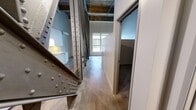 This single-floor loft space makes a striking architectural statement through its bold industrial design. Dominant large-scale steel trusses — featuring riveted connections in both painted and raw finishes — cut dramatically across the open floor plan, serving as the defining visual element of the space. These structural members rise toward exposed timber beam ceilings, reinforcing the building's historic character and providing impressive vertical volume overhead.

The polished concrete floors extend throughout the main living area, lending a sleek, urban quality that complements the raw industrial framework. Natural light enters through a prominent window at the far end of the space, brightening the open layout and highlighting the interplay of textures throughout.

The kitchen area is clearly defined, featuring white cabinetry, a vibrant yellow tile backsplash, and a wall-mounted mini-split HVAC unit — offering a pop of color against the neutral industrial palette. The hallway connects the main area to the bathroom, which includes a clean, functional tub-shower combo with white surround and chrome fixtures.

Overall, the space delivers a cohesive industrial-loft aesthetic, combining raw structural honesty with carefully chosen modern touches, resulting in a compact yet visually dynamic environment full of architectural character.