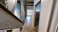 This single-floor loft space makes a striking architectural statement through its bold industrial design. Dominant large-scale steel trusses — featuring riveted connections in both painted and raw finishes — cut dramatically across the open floor plan, serving as the defining visual element of the space. These structural members rise toward exposed timber beam ceilings, reinforcing the building's historic character and providing impressive vertical volume overhead.

The polished concrete floors extend throughout the main living area, lending a sleek, urban quality that complements the raw industrial framework. Natural light enters through a prominent window at the far end of the space, brightening the open layout and highlighting the interplay of textures throughout.

The kitchen area is clearly defined, featuring white cabinetry, a vibrant yellow tile backsplash, and a wall-mounted mini-split HVAC unit — offering a pop of color against the neutral industrial palette. The hallway connects the main area to the bathroom, which includes a clean, functional tub-shower combo with white surround and chrome fixtures.

Overall, the space delivers a cohesive industrial-loft aesthetic, combining raw structural honesty with carefully chosen modern touches, resulting in a compact yet visually dynamic environment full of architectural character.
