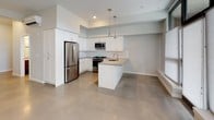 This single-floor unit presents a clean, contemporary design defined by polished concrete floors that flow consistently throughout the space. The open-concept kitchen serves as a central feature, outfitted with white shaker-style cabinetry, a full-height pantry cabinet, stainless steel appliances including a gas range, microwave, refrigerator, and dishwasher, and a peninsula with an integrated sink and neutral-toned countertops. Sleek pendant lighting hangs above the peninsula, adding a modern accent. The adjacent living area is generously proportioned, featuring large windows dressed with light-filtering shades that allow ample natural light to brighten the space. The unit includes two bathrooms: a full bathroom with a soaking tub and shower combination, a gray vanity with a mosaic tile backsplash, and a white countertop; and a secondary bathroom featuring a gray vanity and a separate shower area with grab bars. The bedroom is a well-sized, quiet room with polished concrete floors and a clean, minimal aesthetic. Throughout, the soft gray-white palette and modern finishes create a cohesive, understated ambiance.
