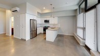 This single-floor unit presents a clean, contemporary design defined by polished concrete floors that flow consistently throughout the space. The open-concept kitchen serves as a central feature, outfitted with white shaker-style cabinetry, a full-height pantry cabinet, stainless steel appliances including a gas range, microwave, refrigerator, and dishwasher, and a peninsula with an integrated sink and neutral-toned countertops. Sleek pendant lighting hangs above the peninsula, adding a modern accent. The adjacent living area is generously proportioned, featuring large windows dressed with light-filtering shades that allow ample natural light to brighten the space. The unit includes two bathrooms: a full bathroom with a soaking tub and shower combination, a gray vanity with a mosaic tile backsplash, and a white countertop; and a secondary bathroom featuring a gray vanity and a separate shower area with grab bars. The bedroom is a well-sized, quiet room with polished concrete floors and a clean, minimal aesthetic. Throughout, the soft gray-white palette and modern finishes create a cohesive, understated ambiance.
