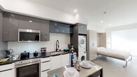 Located on Chandler Boulevard in the North Hollywood neighborhood of Los Angeles, this single-floor studio unit offers a thoughtfully designed open-concept layout that maximizes functionality within a compact footprint. The interior features a clean, contemporary aesthetic with white walls, recessed lighting, and smooth concrete-style flooring that flows consistently throughout the space.

The kitchen is a standout feature, outfitted with high-gloss gray upper cabinetry, crisp white lower cabinets, and dark countertops that create a striking contrast. Stainless steel appliances include a full-size refrigerator, range with oven, and built-in microwave. A textured tile backsplash adds visual depth, while a stainless steel island provides additional prep and dining space. A stacked washer and dryer unit is conveniently integrated directly into the kitchen area.

The sleeping area accommodates a Murphy bed tucked into a warm wood-toned built-in surround, allowing the space to serve dual purposes. A window with a roller shade introduces natural light and connects to a small outdoor-facing area. Adjacent built-in storage with wood cabinetry provides ample organization. The bathroom is clean and minimal, featuring a glass-enclosed shower and neutral finishes. The overall ambiance is modern, efficient, and well-suited to urban living.