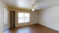 This single-floor residence offers a clean, functional layout designed for comfortable everyday living. The main living area is a standout feature, showcasing warm hardwood-style flooring that extends throughout the space and adds a welcoming, polished feel underfoot. A ceiling fan with an integrated light fixture is centrally mounted above, providing both air circulation and ambient lighting to keep the space comfortable year-round.

Natural light flows in generously through a large window fitted with horizontal blinds, offering views of an outdoor balcony or patio area beyond. The front entry door is positioned conveniently adjacent to this window, creating a bright and open arrival experience. Smooth, neutral-toned walls complement the light color palette of the space, contributing to an airy and versatile atmosphere that can easily accommodate a range of furnishing styles.

A textured ceiling adds a subtle design detail overhead, while clean white baseboards neatly frame the room. A doorway visible to the side hints at additional storage or closet space, enhancing the practicality of the layout. Overall, this space presents a straightforward and well-maintained interior that balances functionality with a bright, inviting character.