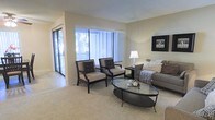 This single-floor residence offers a welcoming and well-appointed living space designed with comfort and functionality in mind. The living room serves as the heart of the home, bathed in natural light streaming through large windows adorned with vertical blinds. A sliding glass door opens to a private balcony, bringing in additional light and providing a seamless connection to the outdoors, with views of surrounding greenery visible through the glass.

The living area features neutral-toned carpeting that adds warmth underfoot, complemented by smooth, light-colored walls that create an airy and open atmosphere. The space is thoughtfully laid out to accommodate both relaxed seating and conversation areas, highlighted by a pair of tufted accent chairs, a cushioned sofa with decorative pillows, and an oval glass-top coffee table that adds a contemporary touch.

A floor lamp provides soft ambient lighting, while framed wall art adds a finishing decorative detail. The overall ambiance is calm, clean, and modern, with a neutral color palette throughout that lends itself to a variety of interior styles. The layout flows naturally, making the space feel open and inviting while maintaining a sense of coziness and warmth.