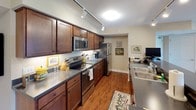 This single-floor residence offers two bedrooms and two full bathrooms within a thoughtfully designed layout. The kitchen is a standout feature, showcasing rich dark wood cabinetry, dark granite-style countertops, stainless steel appliances including a range, built-in microwave, and refrigerator, all complemented by warm hardwood flooring and track lighting overhead. The galley-style configuration maximizes efficiency while maintaining an attractive aesthetic.

The living room provides a relaxed, comfortable atmosphere with plush carpeting, recessed lighting, and a large window fitted with vertical blinds that allows natural light to filter in while offering views of the outdoors. An adjacent dining area features sliding door access and additional natural light, making it well-suited for everyday meals or entertaining.

Both bedrooms are carpeted and generously sized, featuring windows with vertical blinds and ample space for furniture. The primary bathroom includes a wide vanity with an integrated sink, a large mirror, white cabinetry, tile flooring, and a walk-in shower. The second bathroom mirrors a similarly clean, functional design with a single-sink vanity and mirrored medicine cabinet. Warm neutral tones are carried consistently throughout the residence.