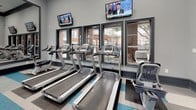 This fitness center is a thoughtfully designed, single-floor facility that offers a comprehensive workout environment for all types of exercise. The space features elevated ceilings with dark crown molding accents, recessed lighting, and ceiling fans that contribute to a comfortable and energizing atmosphere.

The flooring is a distinctive feature, composed of bold striped tiles in teal, gray, and white that add a modern, dynamic aesthetic throughout the room. Large windows along one wall flood the space with natural light and offer views of the surrounding outdoor environment, creating an open and inviting feel. A set of glass-panel double doors provides additional access and visual connection to adjacent areas.

The cardio zone is lined with multiple treadmills positioned directly in front of the windows, accompanied by wall-mounted flat-screen televisions for entertainment during workouts. Elliptical machines and additional cardio equipment are also available throughout the space.

The strength training area is anchored by a collection of cable and resistance machines, as well as bench stations. Full-length mirrors span a large portion of one wall, visually expanding the space and supporting proper form during exercise. A compact storage cabinet and beverage refrigerator offer convenient amenities. The overall design balances functionality with a clean, contemporary style.