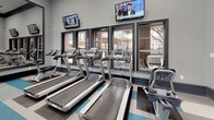 This fitness center is a thoughtfully designed, single-floor facility that offers a comprehensive workout environment for all types of exercise. The space features elevated ceilings with dark crown molding accents, recessed lighting, and ceiling fans that contribute to a comfortable and energizing atmosphere.

The flooring is a distinctive feature, composed of bold striped tiles in teal, gray, and white that add a modern, dynamic aesthetic throughout the room. Large windows along one wall flood the space with natural light and offer views of the surrounding outdoor environment, creating an open and inviting feel. A set of glass-panel double doors provides additional access and visual connection to adjacent areas.

The cardio zone is lined with multiple treadmills positioned directly in front of the windows, accompanied by wall-mounted flat-screen televisions for entertainment during workouts. Elliptical machines and additional cardio equipment are also available throughout the space.

The strength training area is anchored by a collection of cable and resistance machines, as well as bench stations. Full-length mirrors span a large portion of one wall, visually expanding the space and supporting proper form during exercise. A compact storage cabinet and beverage refrigerator offer convenient amenities. The overall design balances functionality with a clean, contemporary style.