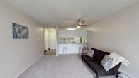 This single-floor space is thoughtfully laid out to maximize functionality and comfort. The living area features soft, light-toned carpeting underfoot and neutral wall tones that create a clean, welcoming ambiance. A ceiling fan with an integrated light fixture anchors the space, providing both airflow and illumination.

The kitchen is positioned adjacent to the living area in an open-concept arrangement, separated by a raised breakfast bar that accommodates seating for two. White cabinetry lines the upper kitchen walls, complemented by a mirrored backsplash that adds a sense of depth and brightness. The kitchen includes standard appliances and provides ample counter space for everyday use.

A hallway visible from the main living area leads to additional rooms, with hard-surface flooring transitioning from the carpeted living space, adding a practical touch near the entryway. The overall layout is compact yet efficient, with a seamless flow between the living and kitchen zones.

The space benefits from consistent overhead lighting and a cohesive neutral color palette throughout, contributing to a bright and airy feel. The open-plan design and functional features make this a practical and comfortable living environment.