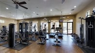 This fitness center offers a comprehensive and well-appointed workout environment designed to meet a variety of exercise needs. The space features warm hardwood-style flooring throughout, creating an inviting atmosphere that complements the functional layout. A generous selection of cardio equipment — including treadmills, ellipticals, and stationary bikes — is arranged in organized rows, each unit equipped with its own personal monitor for entertainment during workouts. Multiple flat-screen TVs are mounted at eye level, ensuring visibility from virtually any point in the room. Large windows and glass doors line the perimeter, flooding the space with abundant natural light and offering pleasant views of the outdoor surroundings. Ceiling fans are suspended throughout, contributing to comfortable airflow during intense sessions. Floor-to-ceiling mirrors on one wall visually expand the space while allowing users to monitor their form. Strength training equipment and free weights are also available, providing a well-rounded fitness experience. Recessed lighting supplements the natural light for consistent illumination throughout the day. The overall ambiance is clean, modern, and energizing — a thoughtfully designed space that supports an active and healthy lifestyle.
