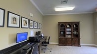 This single-floor business center offers a functional and refined environment designed to meet a variety of professional and personal computing needs. The room is anchored by a long, dark-finished workstation desk that accommodates multiple computer terminals, each equipped with monitors, keyboards, and a printer, providing residents or guests with convenient access to essential office resources.

Overhead, a flush-mounted fluorescent light fixture ensures bright, even illumination throughout the workspace. The warm beige walls are accented by crisp white crown molding, lending the room a polished, traditional character. A series of black-framed architectural photographs lines one wall, adding a tasteful decorative element that enhances the overall aesthetic.

A stately, dark wood bookcase with arched panel detailing stands prominently along the opposite wall, offering generous shelving for books, binders, and decorative objects. Ergonomic rolling chairs provide comfortable seating at the workstations. The carpeted flooring adds warmth underfoot and contributes to a quieter, more focused atmosphere. Together, these elements create a space that balances practicality with classic style, making it a welcoming and well-equipped destination for work or study.