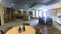 This single-floor communal lobby presents a well-appointed lounge environment designed for comfort and functionality. The space is organized into distinct seating zones, including a casual conversation area furnished with plush sofas and armchairs arranged around a central grouping, as well as additional seating clusters near the windowed perimeter. Large windows fitted with horizontal blinds line the walls, allowing natural light to filter throughout the interior while maintaining privacy. Recessed lighting and flush-mount ceiling fixtures supplement illumination, ensuring the space remains bright and welcoming. The ceiling features subtle tray-style detailing with defined borders, adding architectural interest overhead. A patterned carpet in neutral earth tones covers the floor, contributing texture and visual warmth across the open layout. Decorative touches include tall indoor potted plants, framed wall art, and elegant glass sculptural accents displayed on a granite-topped round table in the foreground. Stone-tile accent walls provide contrast against the neutral color palette. Square columns punctuate the space, contributing to the structured layout while defining separate areas within the open floor plan. Overall, the lobby conveys a professional yet comfortable ambiance suited for gathering and relaxation.