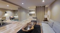 This single-floor space showcases a thoughtfully designed open-concept layout that blends functionality with modern style. The living area is anchored by a striking live-edge wooden coffee table, serving as a natural focal point against the dark area rug and light hardwood flooring that extends throughout the space. Recessed lighting in the smooth ceiling provides consistent, warm illumination across the room. Neutral gray walls create a calm, cohesive backdrop that complements the contemporary furnishings, including a gray sofa and accent chair.

A compact kitchenette is integrated toward the back of the space, featuring white cabinetry, a small sink, and efficient counter space — ideal for light food preparation. A floor-to-ceiling curtain panel in a muted taupe tone offers a flexible room divider, adding texture and softness to the space. A secondary area is partially visible through a structural column, where open shelving and a seating arrangement suggest an additional lounge or reading nook. The overall ambiance is clean, understated, and well-organized, reflecting a modern interior sensibility.