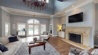This single-floor clubhouse offers a sophisticated and welcoming atmosphere, thoughtfully designed for both relaxation and social gatherings. The main living area commands attention with dramatically soaring tray ceilings adorned by an ornate wrought-iron chandelier, creating a grand yet inviting ambiance. Soft blue-gray walls paired with crisp white crown molding lend the space a refined, cohesive aesthetic throughout.

The lounge area is anchored by a striking stone fireplace with an elegant travertine surround and decorative wrought-iron screen, above which a wall-mounted flat-screen television is positioned. Hardwood-style flooring runs throughout, complemented by plush area rugs and an arrangement of comfortable white upholstered furniture, patterned accent chairs, and rich wood coffee tables.

Large arched French doors and flanking windows flood the interior with natural light while offering direct views of and access to the outdoor pool area. A dedicated seating and dining nook is visible toward the rear of the space, providing additional areas for gathering. Recessed lighting and carefully placed table lamps ensure warm, layered illumination throughout. The overall design balances classic architectural details with comfortable, community-focused furnishings, making this clubhouse a welcoming hub for residents.