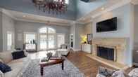 This single-floor clubhouse offers a sophisticated and welcoming atmosphere, thoughtfully designed for both relaxation and social gatherings. The main living area commands attention with dramatically soaring tray ceilings adorned by an ornate wrought-iron chandelier, creating a grand yet inviting ambiance. Soft blue-gray walls paired with crisp white crown molding lend the space a refined, cohesive aesthetic throughout.

The lounge area is anchored by a striking stone fireplace with an elegant travertine surround and decorative wrought-iron screen, above which a wall-mounted flat-screen television is positioned. Hardwood-style flooring runs throughout, complemented by plush area rugs and an arrangement of comfortable white upholstered furniture, patterned accent chairs, and rich wood coffee tables.

Large arched French doors and flanking windows flood the interior with natural light while offering direct views of and access to the outdoor pool area. A dedicated seating and dining nook is visible toward the rear of the space, providing additional areas for gathering. Recessed lighting and carefully placed table lamps ensure warm, layered illumination throughout. The overall design balances classic architectural details with comfortable, community-focused furnishings, making this clubhouse a welcoming hub for residents.
