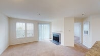 Located at 205 Century Oak in Alexandria, Virginia, this single-floor residence offers a well-designed layout with a seamless flow between its living spaces. The main living area is generously sized, featuring warm neutral carpet, a tile-surround wood-burning fireplace as a central focal point, and large windows fitted with blinds and vertical panel drapes that allow ample natural light. A sliding glass door in the living room provides direct access to the outdoors. Built-in shelving flanks the fireplace wall, offering additional storage and display space. The kitchen is thoughtfully designed with white cabinetry, dark granite-style countertops, a tile backsplash, and white appliances including a range, microwave, and refrigerator. A curved breakfast bar peninsula connects the kitchen to the open carpeted dining and living area, making it ideal for casual entertaining. The bedroom is a comfortable, light-filled space with plush carpet, a ceiling fan with integrated lighting, and a window with an exterior view. The full bathroom features wood-look plank flooring, white cabinetry with a granite-style vanity top, and a tub-shower combination. Throughout the residence, a clean, neutral color palette and consistent finishes create a cohesive and inviting atmosphere.