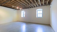 This single-floor space offers an open, loft-style layout that blends raw industrial character with clean, minimalist finishes. The most striking architectural feature is the exposed wooden beam ceiling, which showcases the building's structural framework and adds warmth and texture to the overhead expanse. Track lighting is integrated into the ceiling, providing flexible illumination throughout the space. The polished concrete floors reflect light and contribute to a sense of openness and continuity across the entire floor plan. Two arched windows with dark-framed grid panes punctuate the smooth, light-colored walls, allowing generous amounts of natural light to pour in and brighten the interior. The arch detailing on the windows hints at a historic or character-rich building origin, adding a touch of architectural elegance to the otherwise utilitarian aesthetic. The walls are finished in a clean, neutral tone that complements both the raw ceiling above and the polished floor below. The overall ambiance is spacious, airy, and versatile — well-suited for creative, commercial, or event-based uses that benefit from an open, unobstructed layout with compelling architectural detail.