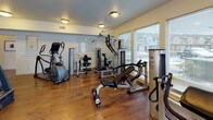 This fitness center is a single-floor facility designed to support a comprehensive workout experience. The space is finished with warm hardwood flooring throughout, providing a clean and durable surface that accommodates a wide range of equipment. Neutral, light-toned walls and flush-mount ceiling fixtures contribute to a bright and functional atmosphere.

The room is generously proportioned, housing an extensive selection of strength-training machines, including cable systems, leg press units, an adjustable bench, and a multi-station cable tower. Cardio equipment includes two treadmills and an elliptical trainer, positioned to take advantage of the natural light that enters through the large windows and glass-paneled exterior door along the front-facing wall. These windows offer views of the surrounding residential environment and allow daylight to fill the space throughout the day.

Additional amenities include a dual water fountain station mounted on the wall, providing convenient hydration access. The layout is well-organized, with clear circulation paths between equipment. The combination of natural light, hardwood flooring, and a diverse equipment selection makes this a practical and inviting fitness facility.