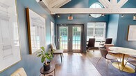 This compact yet thoughtfully designed space makes a strong architectural impression through its soaring vaulted ceiling with exposed white-painted wooden beams and trusses, which lend a sense of volume and openness well beyond its modest footprint. Walls painted in a rich teal-blue create a bold, sophisticated backdrop throughout the interior.

Natural light pours in generously from multiple sources, including large windows with slatted blinds, French doors that open to an outdoor area with a visible pool, a distinctive circular porthole-style window, and a row of clerestory windows set into the roofline. Wood-look tile flooring in warm tones runs throughout, adding warmth and continuity.

A central focal point is a substantial concrete fireplace surround, providing an elegant and grounding architectural element. Frosted glass partition walls divide a dedicated workspace area, outfitted with a built-in marble-top desk and ergonomic seating, offering both privacy and visual openness. A round marble-top table with sculptural seating anchors a central gathering or meeting area. Track lighting and pendant fixtures supplement natural light throughout.

Architectural artwork in gold frames adorns the teal walls, completing the refined, design-forward aesthetic of this versatile and inviting space.