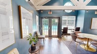 This compact yet thoughtfully designed space makes a strong architectural impression through its soaring vaulted ceiling with exposed white-painted wooden beams and trusses, which lend a sense of volume and openness well beyond its modest footprint. Walls painted in a rich teal-blue create a bold, sophisticated backdrop throughout the interior.

Natural light pours in generously from multiple sources, including large windows with slatted blinds, French doors that open to an outdoor area with a visible pool, a distinctive circular porthole-style window, and a row of clerestory windows set into the roofline. Wood-look tile flooring in warm tones runs throughout, adding warmth and continuity.

A central focal point is a substantial concrete fireplace surround, providing an elegant and grounding architectural element. Frosted glass partition walls divide a dedicated workspace area, outfitted with a built-in marble-top desk and ergonomic seating, offering both privacy and visual openness. A round marble-top table with sculptural seating anchors a central gathering or meeting area. Track lighting and pendant fixtures supplement natural light throughout.

Architectural artwork in gold frames adorns the teal walls, completing the refined, design-forward aesthetic of this versatile and inviting space.