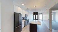 This thoughtfully designed 2-bedroom, 2-bathroom residence offers a seamless open-concept floor plan that connects the kitchen, dining, and living areas into one cohesive space. The contemporary kitchen is a standout feature, outfitted with sleek stainless steel appliances, an electric range, built-in microwave, and a French door refrigerator. Crisp white shaker-style cabinetry extends to the ceiling, maximizing storage, while a two-tone kitchen island with a light quartz countertop provides additional prep space and casual seating. Subtle blue-grey tile backsplash adds a refined accent against the clean white palette. A bold drum pendant light anchors the open dining area, which flows naturally toward the living space. Light-toned hardwood-style flooring runs throughout, lending warmth and continuity to the interior. The living area is bathed in natural light, courtesy of large floor-to-ceiling glass doors that open to an exterior view, enhancing the airy ambiance. Recessed lighting throughout the unit ensures a bright, well-lit environment at any time of day. The overall aesthetic is clean, modern, and move-in ready, making this a highly functional and stylish home.