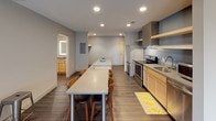 Located at 405 Bridge St NW in Grand Rapids, Michigan, this single-floor unit offers a well-designed, contemporary living space with a functional open-concept layout. The kitchen is a standout feature, equipped with stainless steel appliances including a full-size refrigerator, electric range with a stainless steel hood vent, and microwave. Light maple cabinetry, open wall-mounted shelving with a muted blue accent backdrop, and a light-toned countertop island with built-in electrical outlets complete the kitchen. The elongated island doubles as a dining bar with seating, making it ideal for both meal prep and casual dining. Recessed lighting throughout the kitchen and dining area ensures consistent illumination. The living area is generously proportioned, featuring dark hardwood-style flooring, neutral-toned walls, and multiple windows that allow natural light to filter in alongside views of the surrounding neighborhood. The bathroom provides a practical layout with wood-look vinyl flooring, a tub-shower combination, a louvered closet, and a vanity with warm wood cabinetry. The overall design balances modern finishes with a clean, understated aesthetic throughout.