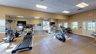 This fitness center is housed in a single-story layout designed to accommodate a comprehensive range of exercise activities. The space features warm beige walls, large-format tile flooring, and a drop ceiling fitted with recessed fluorescent panel lighting that ensures consistent, even illumination throughout. Multiple windows allow natural light to filter in, enhancing the bright and energetic atmosphere.

The cardio zone is outfitted with treadmills, elliptical machines, and a stationary bike, complemented by wall-mounted televisions for entertainment during workouts. A full-length mirror runs along one wall, visually expanding the space and supporting proper form during exercise. The strength training area includes a multi-functional cable machine, a power rack, battle ropes, medicine balls, resistance bands, and a dumbbell rack, offering a versatile range of resistance training options.

Workout reference charts are posted on the walls for user guidance. A water fountain is conveniently located near the entrance, and the space benefits from an open floor plan that allows for flexible use. The clean, functional design and well-maintained equipment make this fitness center a practical and inviting environment for a variety of training needs.