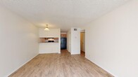 This single-floor unit offers a practical and well-organized layout, accommodating four bedrooms and two full bathrooms. Light wood-style laminate flooring runs consistently throughout the living areas and bedrooms, creating a clean, cohesive aesthetic complemented by neutral white walls and textured ceilings.

The combined kitchen and living room area serves as the heart of the unit. The kitchen features warm-toned wood cabinetry, a black refrigerator and range, a built-in microwave, and a pass-through counter with a granite-style surface that opens to the living area — ideal for casual dining or conversation. A pendant light fixture adds a subtle decorative touch above the pass-through.

The living room is bright and open, with a large window dressed in vertical blinds that invites natural light and outdoor views. Each of the four bedrooms continues the light flooring and neutral palette, with windows fitted with horizontal blinds offering views of surrounding greenery. Bedrooms are served by closets featuring louvered bifold doors and wire shelving systems that maximize storage efficiency.

The two bathrooms are clean and functional, with one featuring a full soaking tub and tiled surround with modern chrome fixtures. A hallway connects the bedroom wing to the main living areas, completing this compact yet capably designed floor plan.
