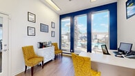This single-floor office suite is a well-appointed, efficiently designed workspace comprising two private offices. The interior is defined by a striking color palette of crisp white walls accented with bold navy blue, creating a professional yet inviting atmosphere. Light wood-style flooring runs throughout, complementing the bright, modern aesthetic.

The reception area greets visitors with an abundance of natural light streaming through large floor-to-ceiling windows framed in navy, offering direct views of an outdoor pool area. Vibrant yellow upholstered chairs and a white decorative cabinet furnish this welcoming space, while framed informational displays line the walls.

The two private offices are separated by sleek glass partition walls, lending an open, transparent feel while maintaining acoustic separation. Each office is equipped with a ceiling fan, ample desk space, and generous window access that fills the rooms with natural light and outdoor views. The consistent use of yellow accent seating and white work surfaces ties the design language together cohesively across both offices.

Overall, the suite strikes a balance between functionality and style, offering a bright, energetic work environment suited for client-facing operations, with thoughtful design details and a warm, professional ambiance throughout.