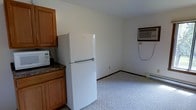 This single-floor space offers a straightforward and functional layout designed for practical everyday living. The kitchen area is outfitted with warm honey-toned wooden cabinetry providing ample storage, a dark granite-style countertop, and a full-size white Whirlpool top-freezer refrigerator. A microwave is also present on the counter, rounding out the essential appliances. The kitchen transitions seamlessly into an open living area, which features light-colored carpeting that contributes to a clean and neutral aesthetic throughout. The walls are finished in a simple off-white tone, giving the space a bright and airy feel. A wall-mounted air conditioning unit is installed in the living area, ensuring climate comfort during warmer months, while a baseboard heater along the wall provides warmth during cooler seasons. The overall design is modest and utilitarian, prioritizing functionality and ease of use. The open-concept flow between the kitchen and living areas makes the space feel cohesive, while the neutral finishes offer flexibility for personalization. This space is well-suited for someone seeking a no-frills, move-in-ready environment with all the core comforts accounted for.