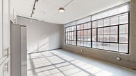 This single-floor space showcases an industrial loft aesthetic, blending raw architectural elements with modern finishes throughout. The main open area is defined by a striking wall of large, multi-pane factory-style windows fitted with black steel frames, flooding the interior with abundant natural light and offering direct views of the surrounding urban streetscape. Polished concrete floors run throughout, complementing the exposed ductwork and track lighting visible along the ceiling.

The kitchen is well-appointed with white cabinetry, a white subway tile backsplash, dark countertops, and a full suite of stainless steel appliances including a refrigerator with ice maker, range, microwave, and dishwasher. Additional upper and lower cabinetry provides generous storage.

Two hallways connect the various areas of the space. One hallway features a bold dark accent wall, creating a strong visual contrast against the lighter surroundings, while exposed ductwork overhead reinforces the industrial character. A stacked washer and dryer unit is also accommodated within the layout.

The bathroom is finished with large-format tile flooring, a vanity with a stone-top sink, a framed mirror, and a wall-mounted grab bar, reflecting a clean and functional design. A separate smaller room offers additional flexible space with concrete floors and neutral walls.