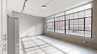This single-floor space showcases an industrial loft aesthetic, blending raw architectural elements with modern finishes throughout. The main open area is defined by a striking wall of large, multi-pane factory-style windows fitted with black steel frames, flooding the interior with abundant natural light and offering direct views of the surrounding urban streetscape. Polished concrete floors run throughout, complementing the exposed ductwork and track lighting visible along the ceiling.

The kitchen is well-appointed with white cabinetry, a white subway tile backsplash, dark countertops, and a full suite of stainless steel appliances including a refrigerator with ice maker, range, microwave, and dishwasher. Additional upper and lower cabinetry provides generous storage.

Two hallways connect the various areas of the space. One hallway features a bold dark accent wall, creating a strong visual contrast against the lighter surroundings, while exposed ductwork overhead reinforces the industrial character. A stacked washer and dryer unit is also accommodated within the layout.

The bathroom is finished with large-format tile flooring, a vanity with a stone-top sink, a framed mirror, and a wall-mounted grab bar, reflecting a clean and functional design. A separate smaller room offers additional flexible space with concrete floors and neutral walls.