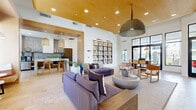 Located in Goodyear, Arizona, this single-floor space offers a thoughtfully designed open-concept layout that seamlessly connects a spacious living room and a well-appointed kitchen. The living room commands attention with soaring ceilings accented by warm wood paneling and recessed lighting, creating an inviting, airy atmosphere. A striking floor-to-ceiling marble fireplace wall, topped by a wall-mounted television, anchors the room, while oversized windows and glass doors flood the interior with natural light and offer views of the surrounding outdoor landscape. Deep-seated sectional sofas in a rich purple hue, natural wood accent furniture, and a sculptural concrete coffee table lend the space a contemporary yet comfortable character. Woven pendant light fixtures add warmth and texture overhead. Intimate nook seating areas lined with green stone tile provide cozy retreats within the larger room, alongside a full-wall built-in bookcase that serves as a bold design statement. The kitchen features dark stone countertops, warm wood cabinetry, and a mosaic tile backsplash. A generous concrete-topped island with bar seating and woven pendant lights overhead makes it ideal for casual dining and entertaining. Large-format tile flooring flows consistently throughout, unifying the entire space with a clean, polished aesthetic.