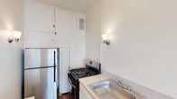 Unit 309 is a well-appointed single-floor residence that blends classic architectural details with practical living spaces. The kitchen is efficiently designed, featuring stainless steel appliances including a full-size refrigerator and a gas range, ample white cabinetry extending to the ceiling for generous storage, a stainless steel sink, and a wall-mounted sconce for warm accent lighting. Rich hardwood flooring anchors the kitchen and connecting areas, lending warmth throughout. The living and bedroom areas are finished with plush carpeting and feature elegant chair-rail molding along the walls, adding a timeless, classic character. Ceiling fans with light fixtures are present in the carpeted rooms, providing both comfort and illumination. One bedroom offers direct access to a bathroom en suite and is brightened by a window with views of the surrounding neighborhood. A dedicated closet with a built-in rod provides practical storage. The bathroom is appointed with classic white subway tile surrounding a full soaking tub and shower combination, complemented by a window that allows natural light to fill the space. A warm-toned wooden entry door adds a distinctive, traditional touch to the unit's overall aesthetic. The thoughtful layout and classic finishes make this a comfortable and functional residence.