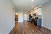 Located at 1409 E Meyer Blvd in Kansas City, Missouri, this single-floor unit presents a thoughtfully designed contemporary layout with a cohesive aesthetic carried throughout each space. The open living and kitchen area serves as the heart of the unit, where rich, dark hardwood-style flooring extends across the main living zones, complementing the neutral gray walls and white trim for a clean, modern look.

The kitchen is well-appointed with a full suite of stainless steel appliances, including a refrigerator, electric range, built-in microwave, and dishwasher. Dark wood cabinetry on the lower portion contrasts neatly with bright white upper cabinets, while granite countertops and a stainless steel sink add a polished finish. Recessed lighting illuminates the space effectively. A dedicated closet adjacent to the kitchen houses a stacked washer and dryer, providing the convenience of in-unit laundry.

The bedroom is carpeted for comfort and benefits from a window that brings in natural light and outdoor views, paired with a ceiling fan overhead. Access to a closet and the bathroom is conveniently located within the bedroom area. The bathroom features subway tile in the shower, light flooring, and a straightforward, functional design with neutral tones throughout.