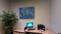 This single-floor office space is designed with productivity and functionality in mind. The workspace centers around a light wood desk that provides ample surface area, accommodating a desktop computer with a monitor, keyboard, and mouse, alongside a multifunction printer — everything needed for day-to-day office operations. The neutral, muted wall tones create a calm and focused atmosphere, while a large vintage-style world map mounted prominently above the workstation serves as both a decorative focal point and an inspiring visual element. A lush green indoor plant is visible to one side, adding a touch of nature and warmth to the otherwise professional environment. The overall ambiance is clean, organized, and purposeful, making it well-suited for administrative or professional work. The understated color palette and practical furnishings come together to create a workspace that is both efficient and visually comfortable for extended use.