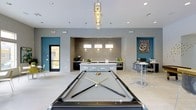 This single-floor amenity space is thoughtfully designed to balance recreation, socializing, and productivity in a contemporary setting. At the heart of the room sits a full-size pool table beneath a sleek, geometric pendant light fixture, serving as the focal point of the open lounge area. The light tile flooring and neutral gray walls are accented by bold teal feature walls, adding a vibrant pop of color to the space.

Toward the back, a stylish kitchenette bar area is framed by a gray tile accent wall and illuminated by a trio of hanging pendant lights. The white countertop bar is lined with modern bar stools and flanked by a coffee station and small appliances, creating a convenient refreshment hub for guests.

To the right, a dedicated "Print & Connect" zone offers high-top tables and workstations, making the space functional for residents needing quick access to business services. Recessed lighting throughout ensures the entire area is well-lit, while large windows and a glass-panel door allow natural light to filter in, contributing to an open and welcoming ambiance. The overall design merges modern aesthetics with practical amenities, making it an ideal communal gathering space.