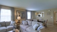 This single-floor space offers a thoughtfully designed living area that blends comfort with a calm, neutral aesthetic. The living room is anchored by plush, light-colored sofas adorned with blue and white decorative pillows, creating an inviting and relaxed atmosphere. Warm beige walls provide a cohesive backdrop throughout, complemented by soft grey curtains framing the windows, which allow natural light to filter into the space.

A framed abstract artwork in earthy tones adds visual interest to the main wall, while a classic table lamp with a bronze base contributes warm ambient lighting. Decorative accents, including a lantern on the floor and small tabletop accessories, lend a curated, homey feel.

A large wall-mounted mirror near the dining area reflects natural light and creates a sense of added depth. The dining area, partially visible beyond the living space, features a dark wood table with upholstered seating, completing the open-concept layout. The overall ambiance is serene and welcoming, with a consistent soft colour palette tying the spaces together seamlessly.