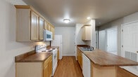 This well-organized 2-bedroom, 1-bathroom single-floor residence offers a practical and comfortable layout. The living room serves as the central gathering space, featuring warm wood-style laminate flooring, a ceiling fan with light fixture, and a large sliding glass door dressed with vertical blinds that allows natural light to fill the room. A diagonal architectural beam accent adds a subtle design element to the ceiling. The galley-style kitchen is efficiently laid out with light wood cabinetry featuring decorative raised panel details, dark laminate countertops, white appliances including a range, over-the-range microwave, refrigerator, and dishwasher, along with a double-basin sink. Wood-style flooring continues throughout the kitchen. The primary bedroom offers a ceiling fan, neutral carpet, and a walk-in closet equipped with wire shelving for organized storage. The secondary bedroom also features wood-style flooring and a ceiling fan. The full bathroom includes a single-sink vanity with warm wood cabinetry, laminate countertop, a tub/shower combination, and wood-style flooring. Neutral gray walls and white trim throughout create a clean, cohesive aesthetic across the entire space.