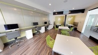 This contemporary multi-story residential community presents a well-designed mix of amenity spaces crafted for both productivity and relaxation. The interior business lounge is a standout feature, combining a dedicated workstation area and a casual dining and lounge zone. The workstation wall accommodates multiple computer terminals along a sleek built-in desk with ample storage below, all set against a frosted glass panel backdrop with lime green accent detailing. Vibrant, lattice-patterned green chairs surround crisp white tables throughout, while warm wood-look flooring and recessed lighting create a polished, energetic atmosphere. Intimate booth-style seating nooks with yellow-green accent walls and sculptural pendant lighting add character to the lounge area. Floor-to-ceiling glass walls connect the interior space seamlessly to the outdoors, flooding the room with natural light and views of the courtyard. A wall-mounted flat-screen display adds a media element to the space. The outdoor courtyard is furnished with comfortable lounge chairs, dining tables, and an outdoor grill, providing a welcoming environment for gathering. The building exterior showcases a multi-story facade with balconies, large windows, and a well-maintained streetscape. A covered parking structure with organized stalls completes the community's practical offerings.