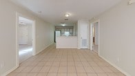 This single-floor residence offers a practical and accessible layout designed for comfortable everyday living. The main living area is finished with light-toned ceramic tile flooring and features a ceiling fan, a front entry door, and a window that allows natural light to fill the space. The kitchen is positioned at the rear of the living area and is defined by a peninsula counter that creates a natural divider. It is equipped with white appliances including a range and refrigerator, complemented by taupe cabinetry and a built-in pantry unit. A set of double panel doors provides additional storage within the kitchen. The bedrooms are carpeted and feature ceiling fans with light fixtures, closet storage, and windows fitted with blinds that invite natural light. The primary bedroom is notably spacious. The bathroom is thoughtfully designed with accessibility in mind, featuring a wide, open roll-in shower with a grab bar, a generous vanity with an open knee space beneath a large mirror illuminated by a Hollywood-style light bar, and tile flooring. The neutral color palette of warm beige and white tones carried throughout the space creates a clean, cohesive, and inviting atmosphere.