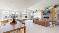 This generously sized single-floor community amenity space is designed to impress and invite. The open layout is anchored by soaring double-height ceilings and an expansive wall of floor-to-ceiling windows and glass doors that flood the interior with natural light and offer views of the outdoor pool area beyond.

The lounge area serves as the heart of the space, featuring a warm, contemporary design highlighted by a dramatic hand-painted botanical mural that sweeps across one wall and up toward the ceiling. A wood-paneled accent wall houses a wall-mounted flat-screen TV above a sleek marble fireplace, creating a refined focal point. Leather sofas, accent chairs, and a patterned area rug complete the comfortable seating arrangement, while a geometric gold pendant light adds an elegant touch overhead.

A dedicated game zone offers a shuffleboard table and casual high-top seating, perfect for social gatherings. Adjacent to the lounge, a well-appointed kitchen and dining area features warm wood cabinetry, open shelving, a marble-tiled backsplash, and stainless steel appliances. A large communal dining table with modern wood-and-leather chairs provides ample space for shared meals or group activities. Curated wall art and decorative accessories throughout contribute to the space's cohesive, modern-coastal aesthetic. Large-format white tile flooring ties the entire space together seamlessly.