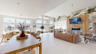 This generously sized single-floor community amenity space is designed to impress and invite. The open layout is anchored by soaring double-height ceilings and an expansive wall of floor-to-ceiling windows and glass doors that flood the interior with natural light and offer views of the outdoor pool area beyond.

The lounge area serves as the heart of the space, featuring a warm, contemporary design highlighted by a dramatic hand-painted botanical mural that sweeps across one wall and up toward the ceiling. A wood-paneled accent wall houses a wall-mounted flat-screen TV above a sleek marble fireplace, creating a refined focal point. Leather sofas, accent chairs, and a patterned area rug complete the comfortable seating arrangement, while a geometric gold pendant light adds an elegant touch overhead.

A dedicated game zone offers a shuffleboard table and casual high-top seating, perfect for social gatherings. Adjacent to the lounge, a well-appointed kitchen and dining area features warm wood cabinetry, open shelving, a marble-tiled backsplash, and stainless steel appliances. A large communal dining table with modern wood-and-leather chairs provides ample space for shared meals or group activities. Curated wall art and decorative accessories throughout contribute to the space's cohesive, modern-coastal aesthetic. Large-format white tile flooring ties the entire space together seamlessly.